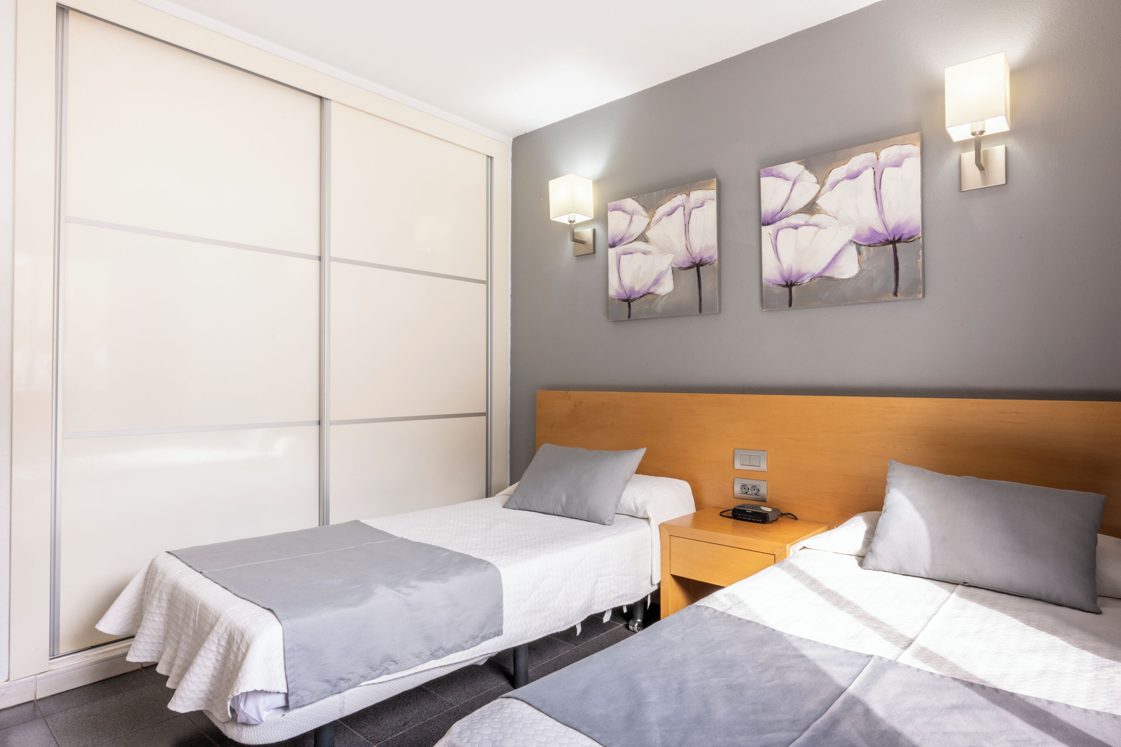Hotellihuone, jossa on kaksi sänkyä, puinen pääty, yöpöytä ja seinään kiinnitetyt valaisimet.