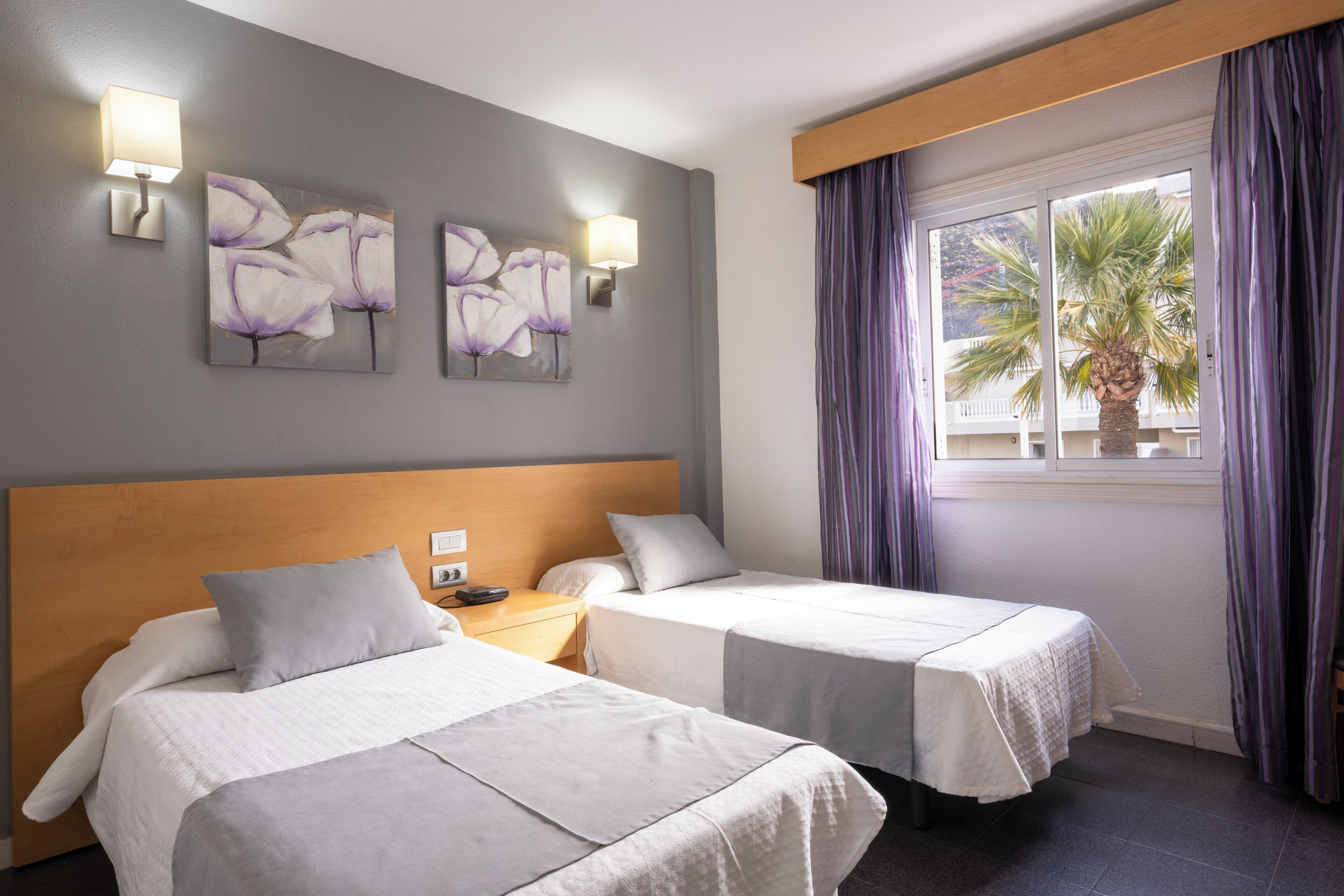 A hotel room with two beds, a wooden headboard, a nightstand, two wall-mounted lamps, and a window with curtains.