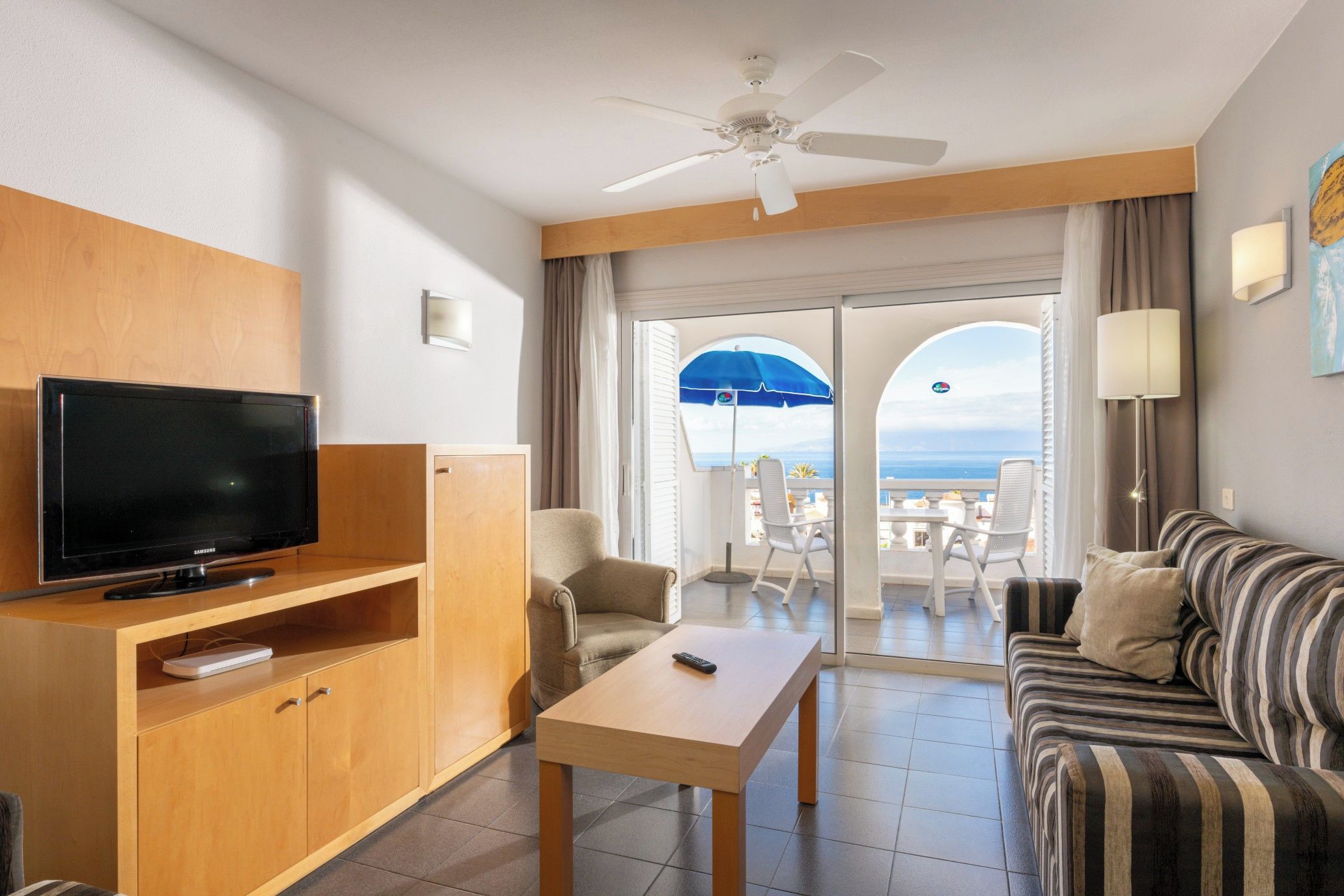 A hotel room with a TV, a sofa, a coffee table, a ceiling fan, and a balcony with a view.