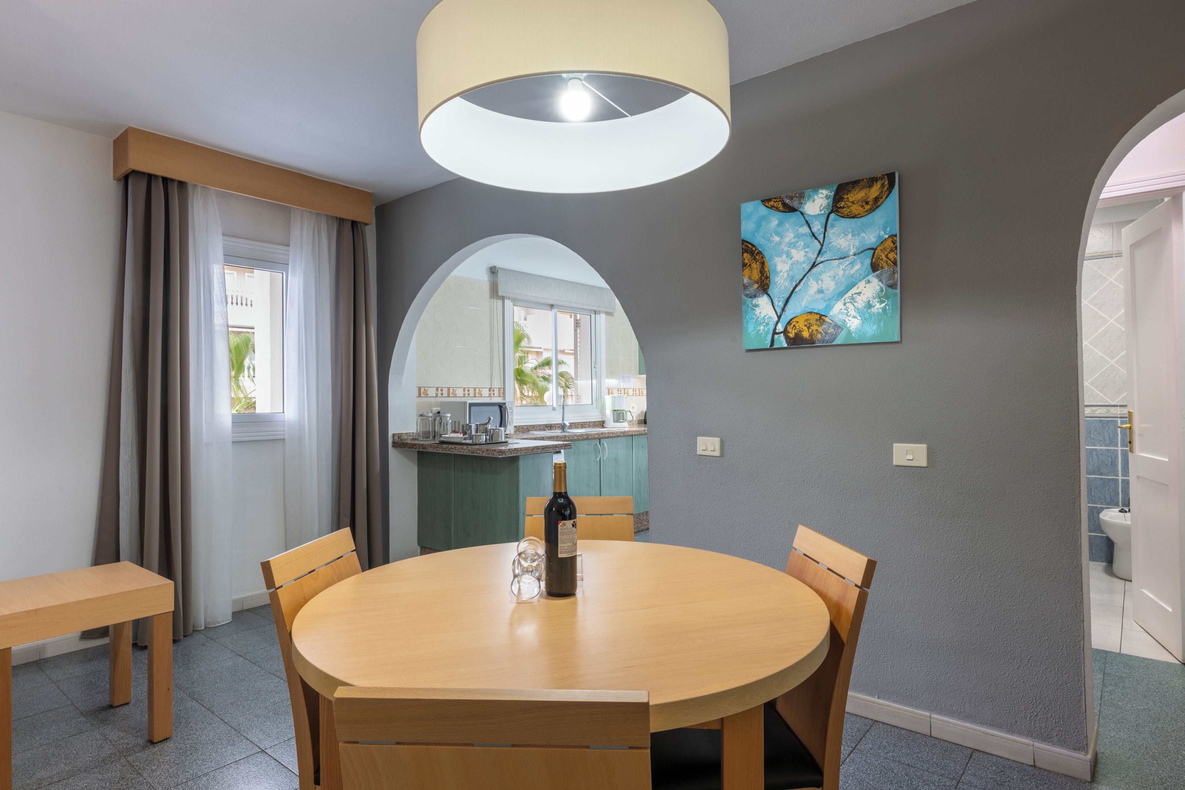 Een eethoek met een ronde tafel, stoelen, een fles wijn, een schilderij en uitzicht op de keuken en het balkon.