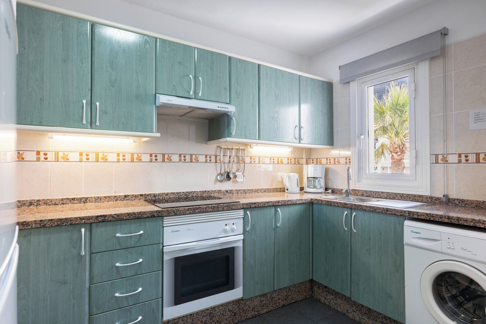 Una cocina con gabinetes verde azulado, horno empotrado, lavadora y ventana con vista a palmeras.