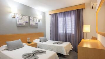 Habitación de hotel con dos camas, una mesita de noche, un escritorio, una lámpara y un ventanal con cortinas.