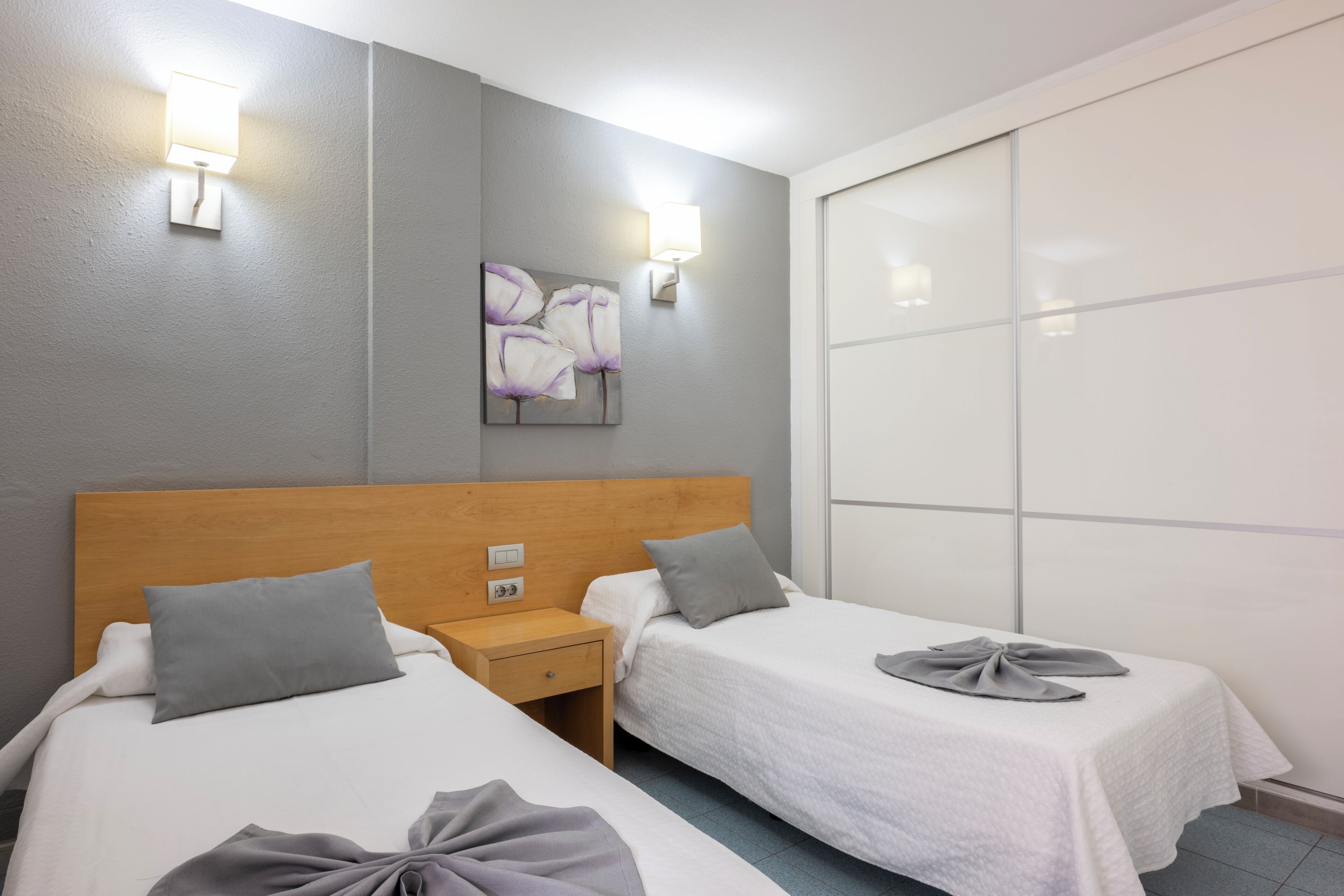Habitación de hotel con dos camas, cabecero de madera, mesita de noche, lámpara de pared y un ventanal grande.