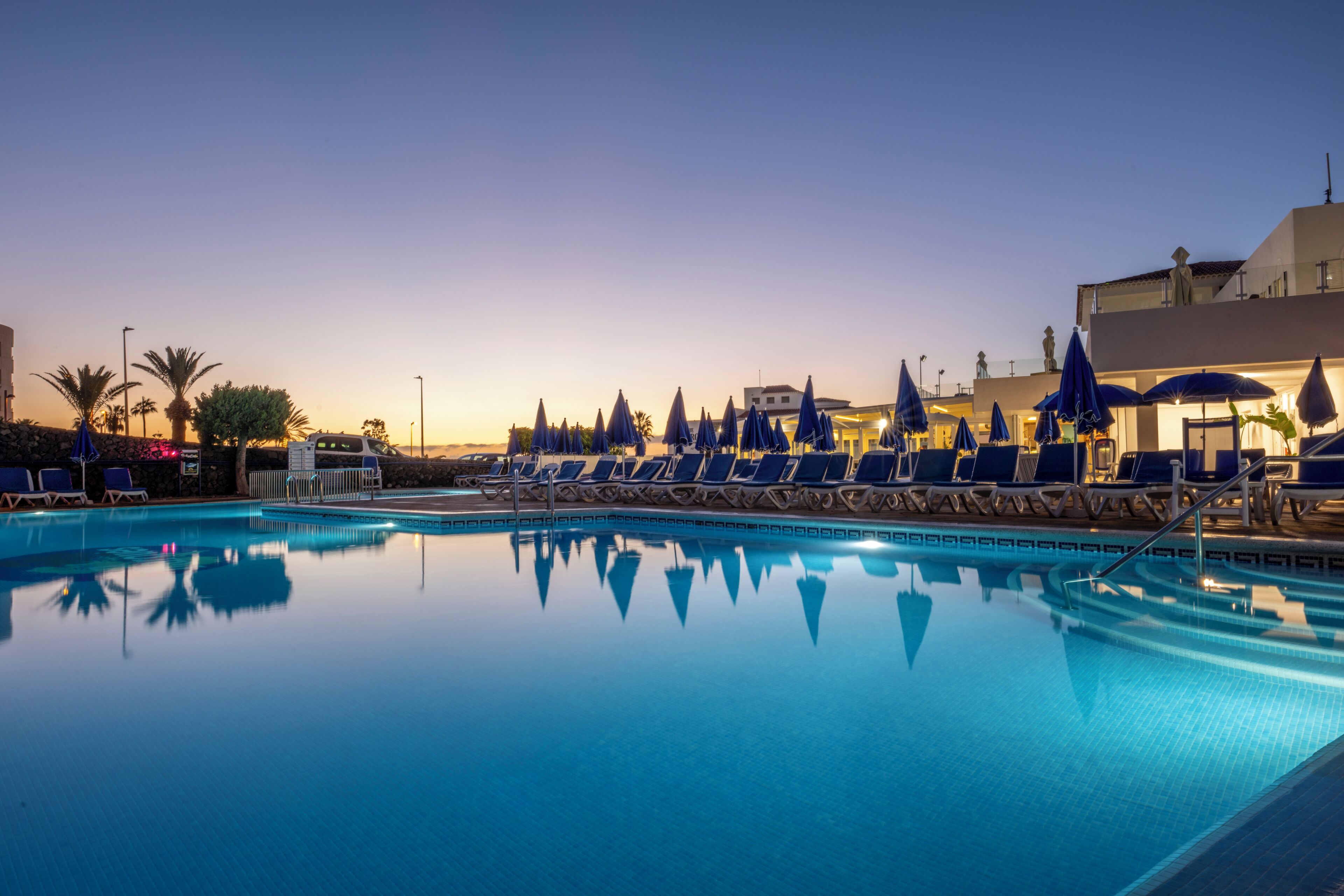 A swimming pool with lounge chairs and umbrellas, surrounded by palm trees and buildings.