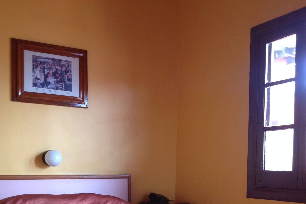 Una habitación con una cama, una mesita de noche, una lámpara, un cuadro en la pared y una ventana.