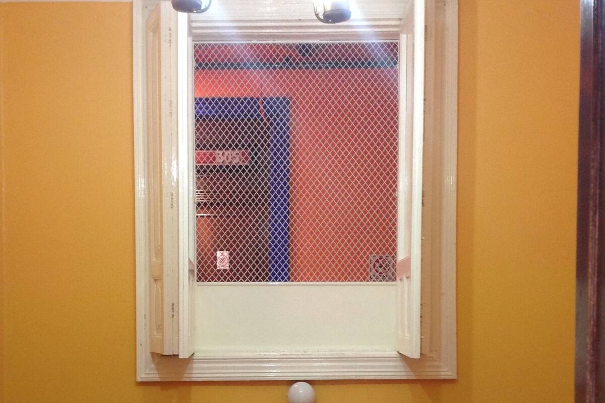 Una habitación pequeña con dos camas, una ventana con mosquitero, una mesita de noche y una lámpara en el techo.