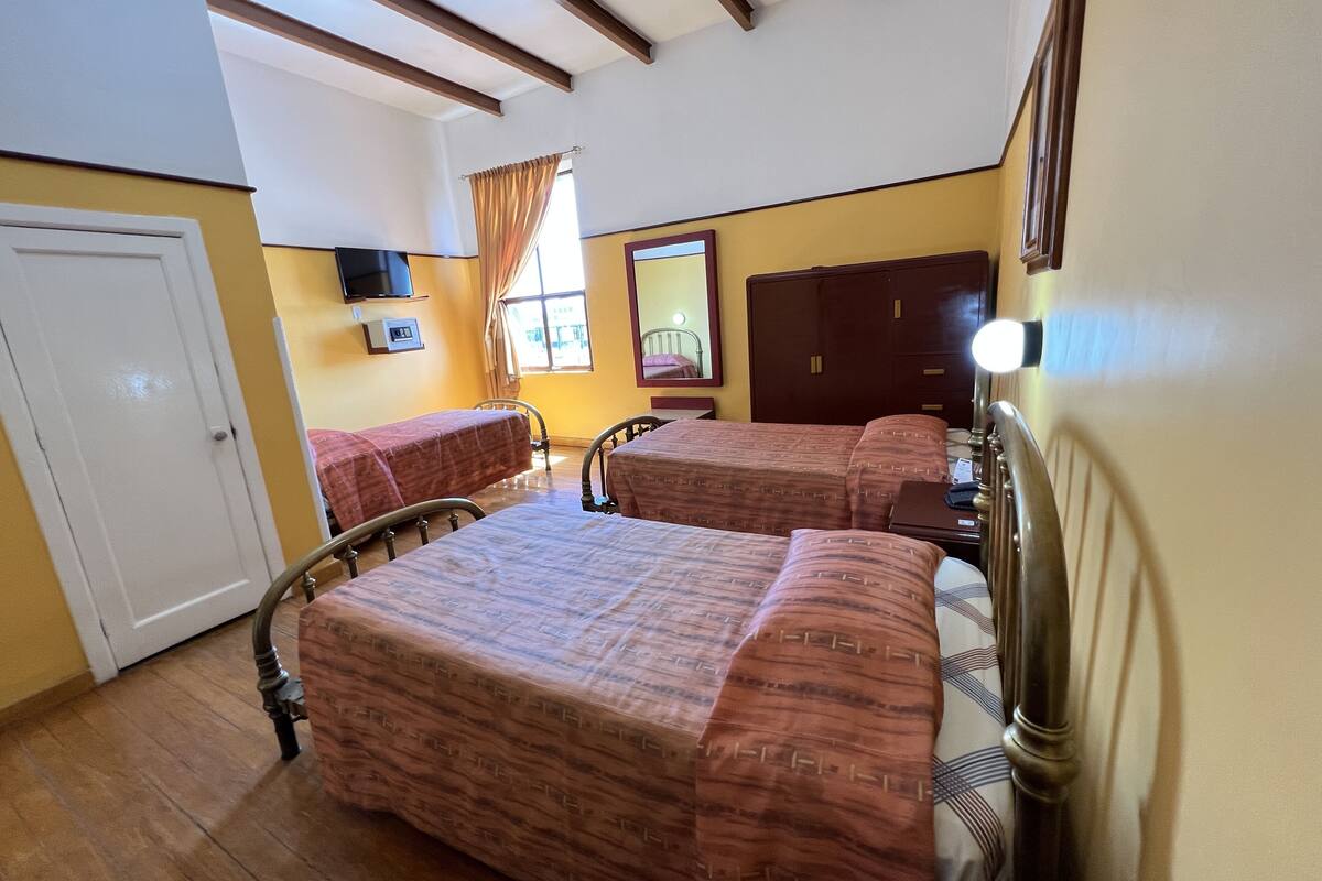 Una habitación con dos camas, techo de madera, ventana con cortinas, un espejo y un televisor en la pared.