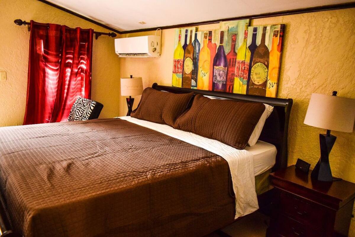 A hotel room with a large bed, red curtains, a wall art of wine bottles, and a nightstand with a lamp.