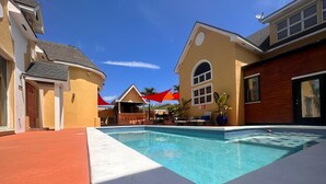 Et poolområde med en klar, blå pool, omgivet af bygninger med gul og rød facade, og en rød parasol, der giver skygge.