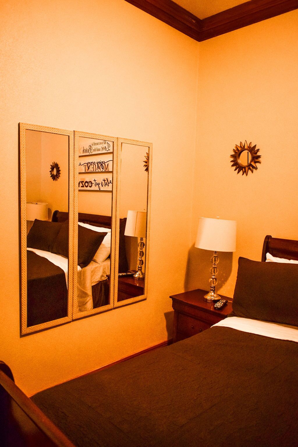 A hotel room with a large mirror, a bed, a nightstand with a lamp, and a wall decoration.