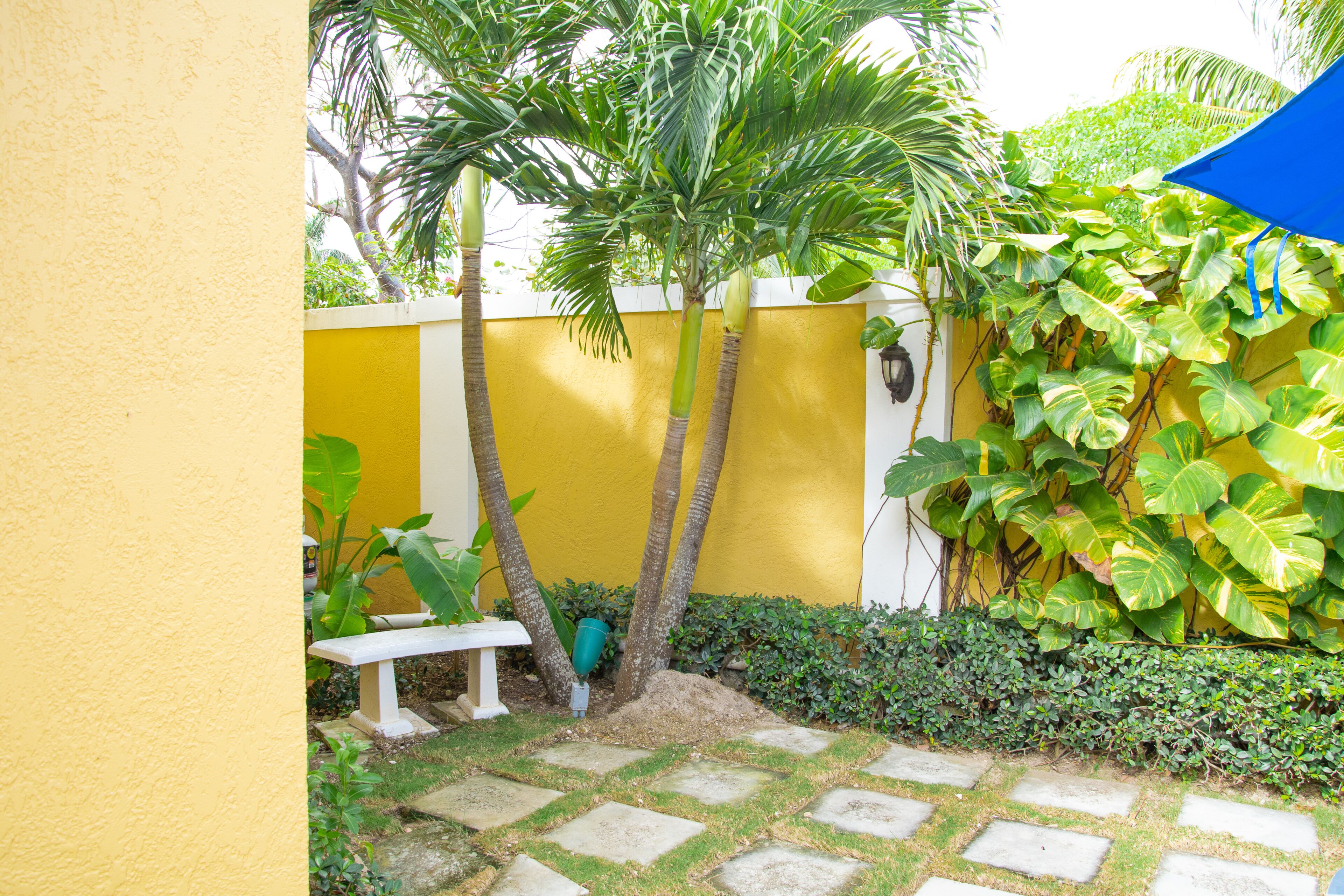 A courtyard with a yellow wall, a white bench, and a blue umbrella.