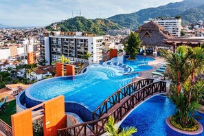 A resort with a large swimming pool, a bridge, and a view of the city and hills in the background.