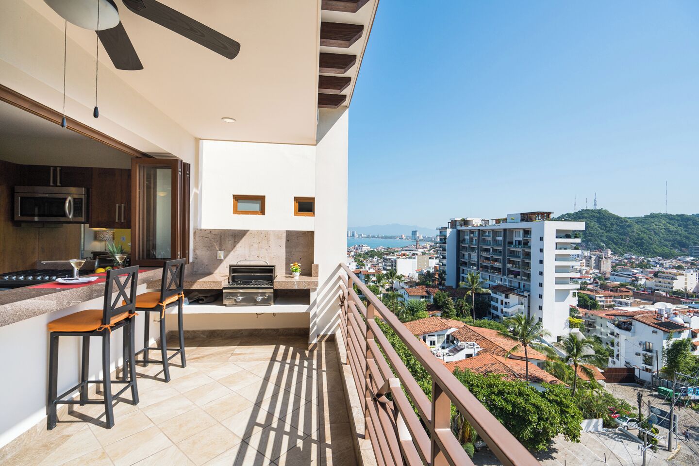 A balcony with a kitchen area, including a grill and bar stools, overlooking a cityscape with buildings and greenery.