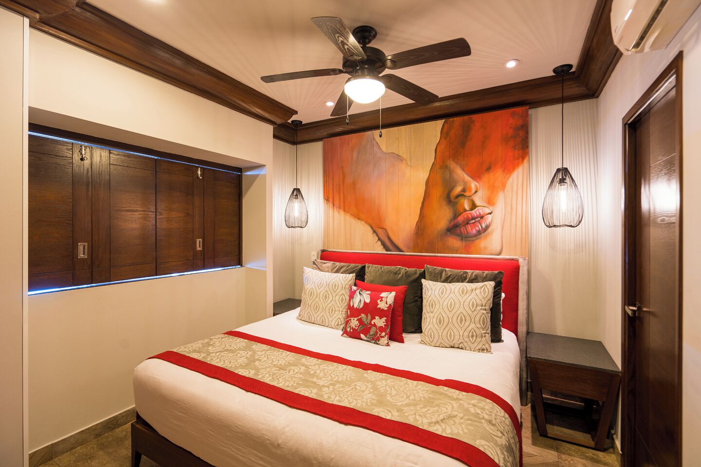 Een slaapkamer met een groot bed, een houten kledingkast, plafondventilator, hanglampen en een muurschildering.