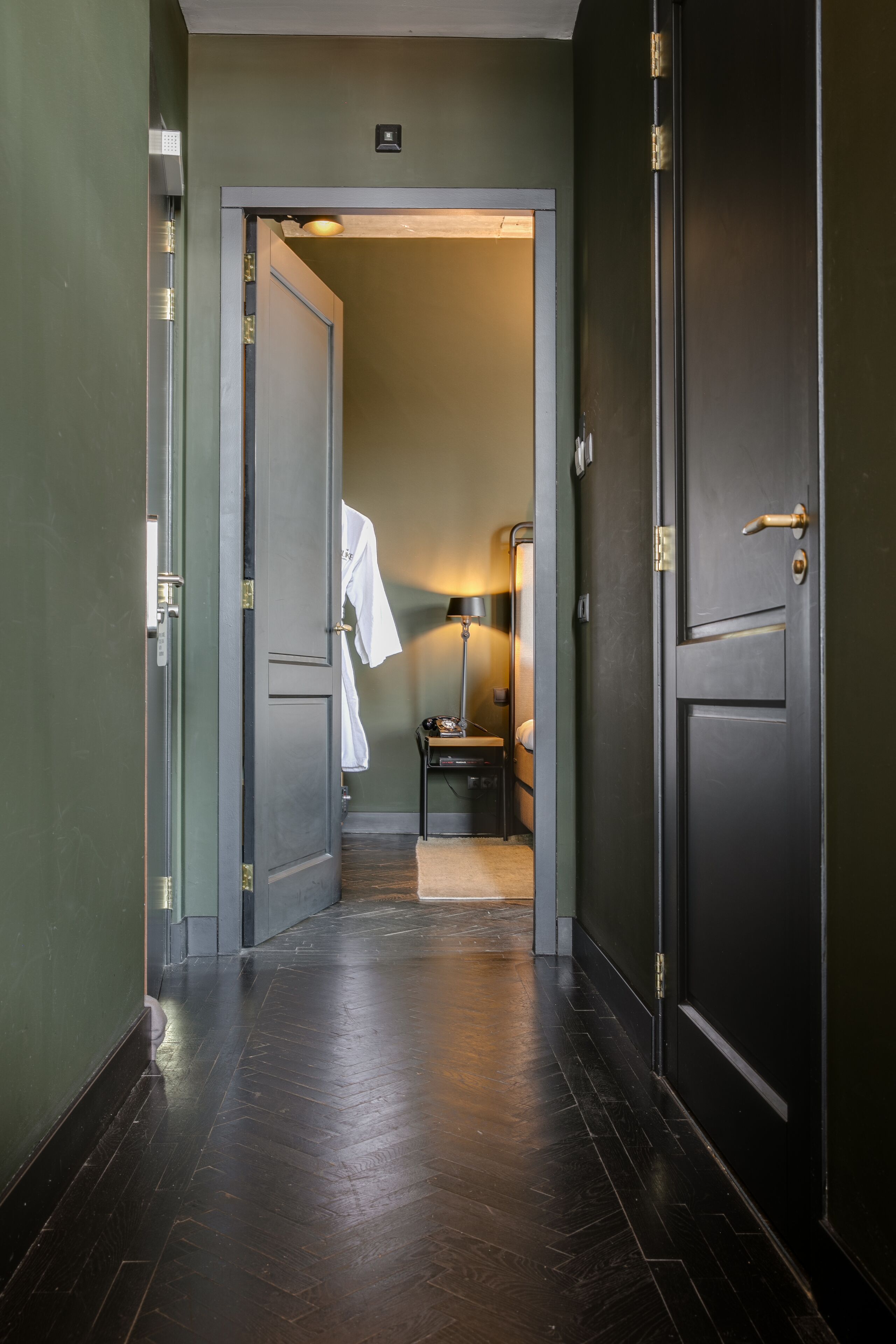 A hallway with dark wooden flooring, a white robe hanging on a door, and a small table with a lamp.