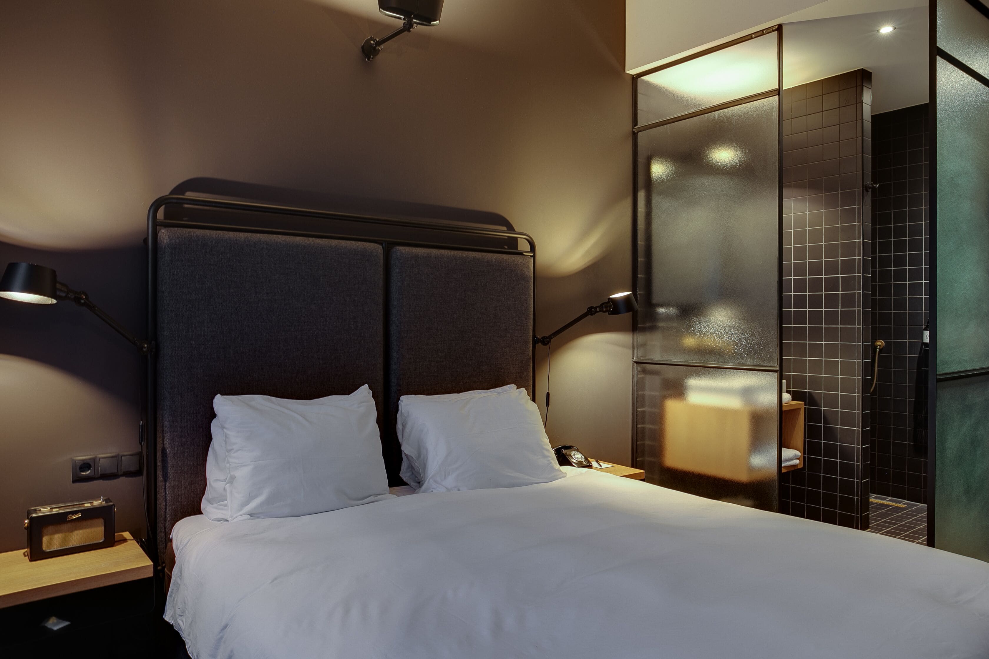 Habitación de hotel moderna con una cama grande, lámparas de noche, una mesita de noche y un área de ducha con mampara de vidrio.