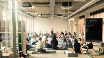 Un amplio estudio de yoga con participantes sentados sobre esterillas, un cartel grande anunciando clases gratuitas, y un techo visible con conductos.