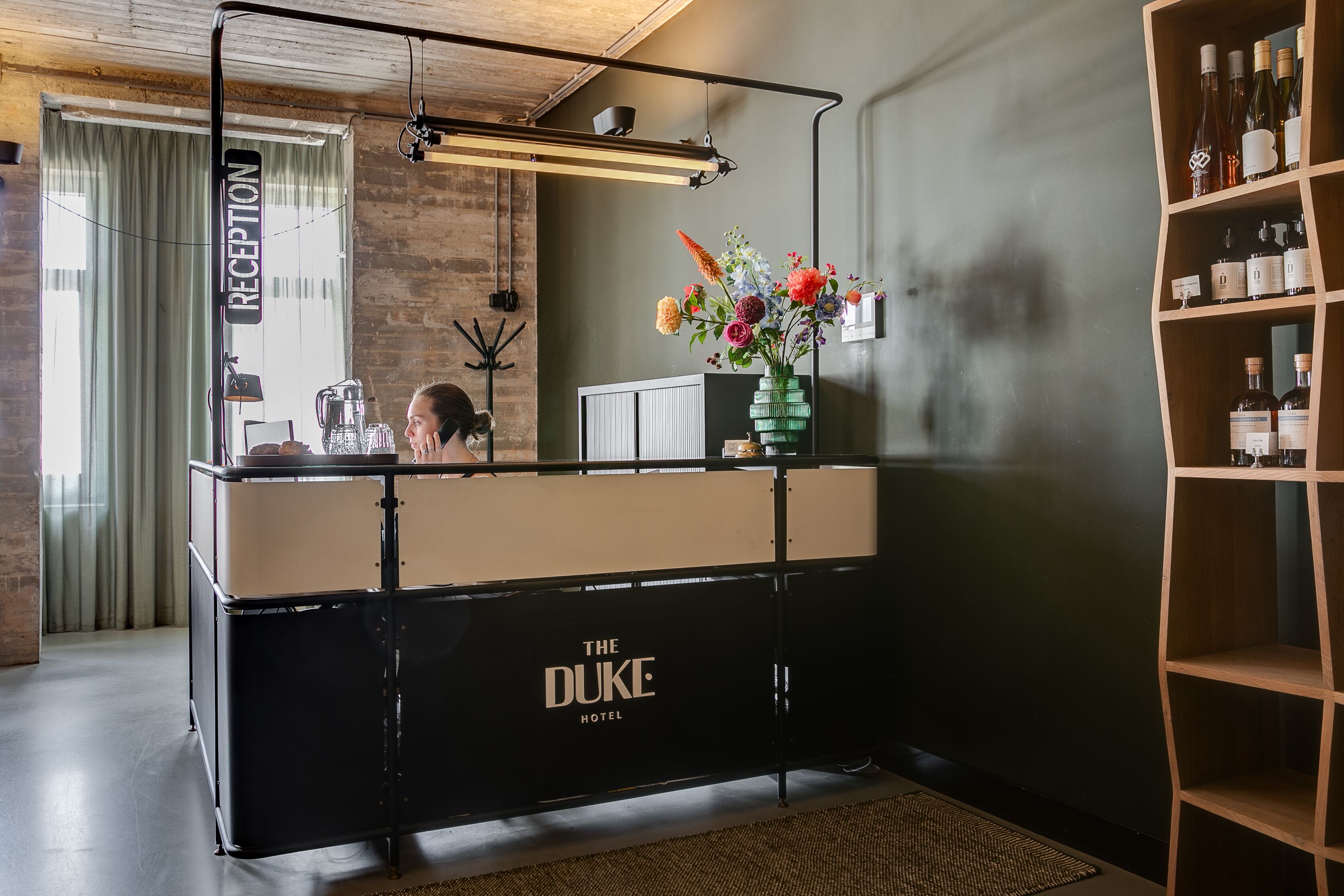 Vastaanottoalue, jossa on musta vastaanottotiski, jossa lukee ”The Duke Hotel”, peili, maljakko kukkineen ja hylly pulloja varten.