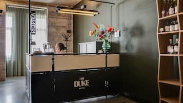 Zona de recepción con mostrador negro con la leyenda "The Duke Hotel", un espejo, un florero con flores y una estantería con botellas.