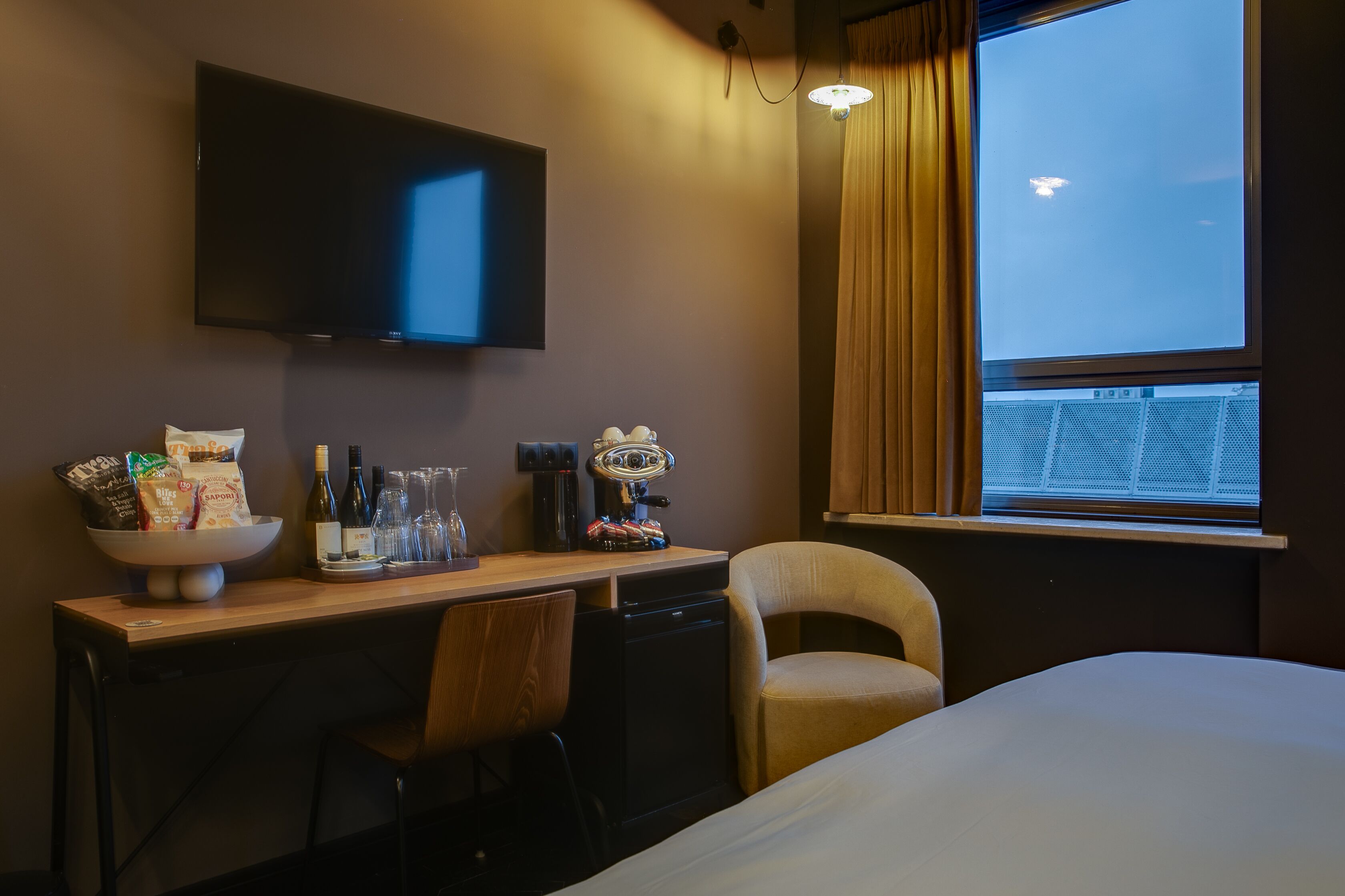 A hotel room with a bed, a desk with a bowl of snacks, a coffee maker, and a television on the wall.