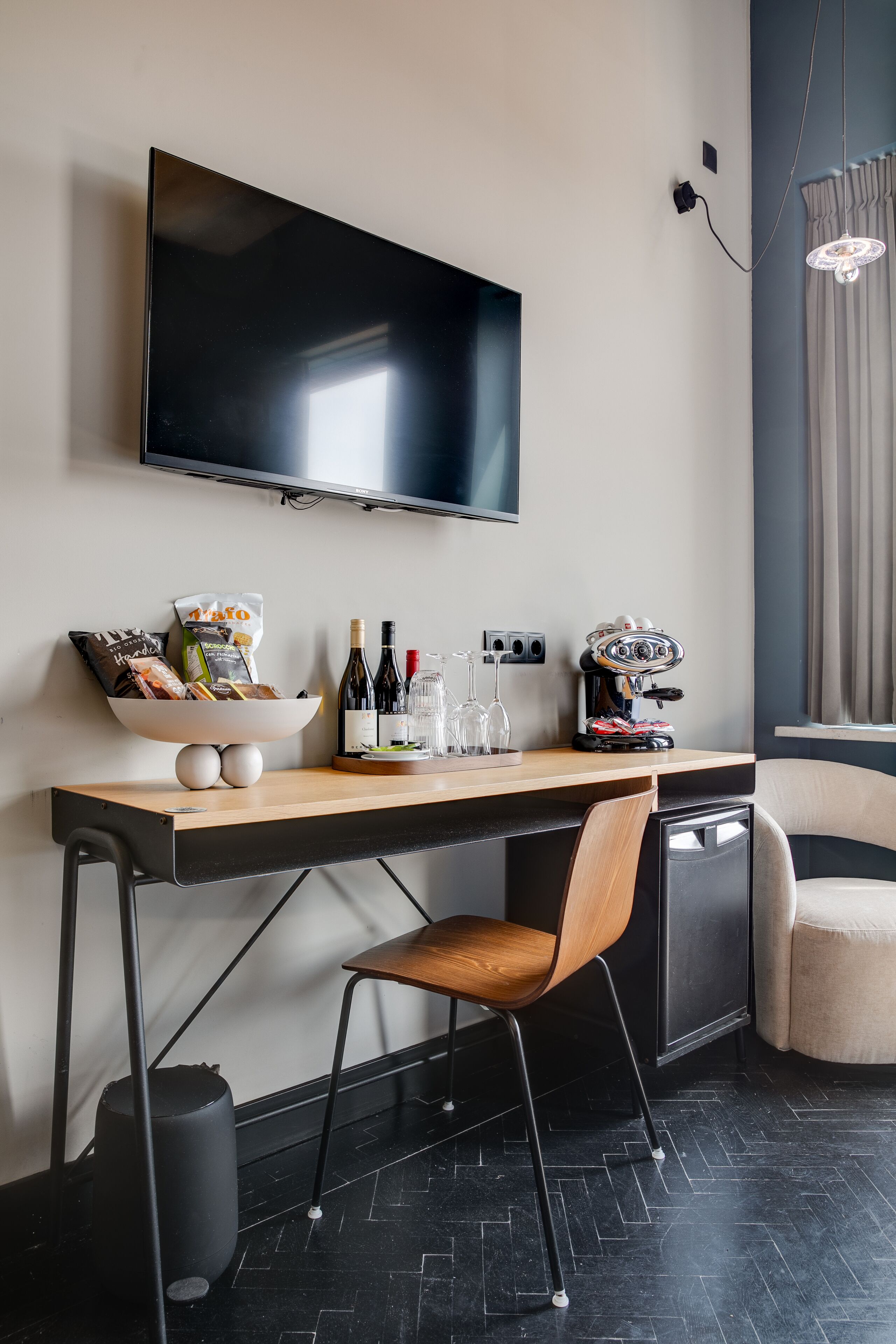 Une chambre d’hôtel moderne équipée d’un téléviseur à écran plat, d’un bureau en bois avec un panier de collations, de bouteilles de vin et d’une machine à café.