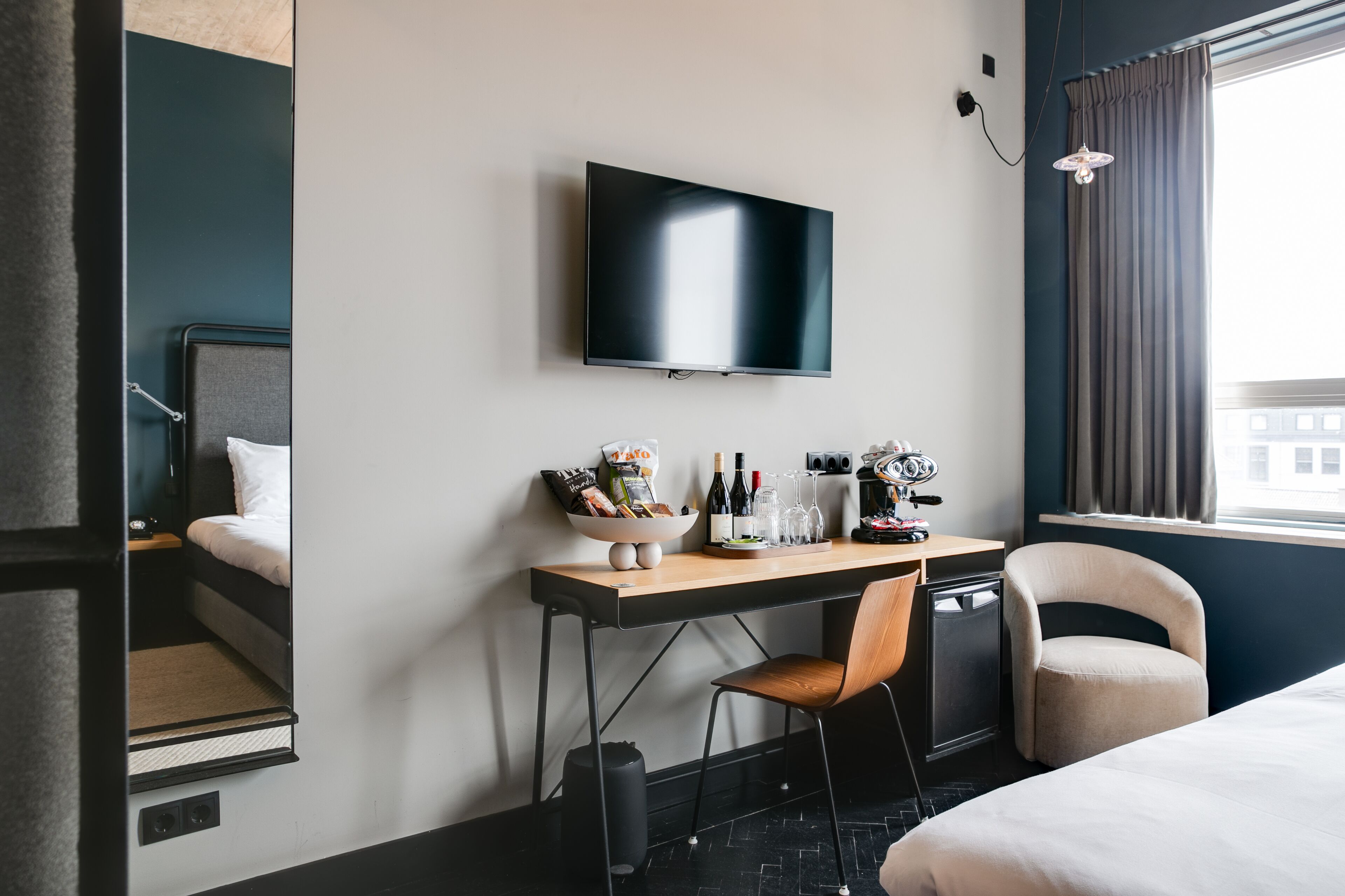 Habitación de hotel con cama, escritorio con silla, televisor montado en la pared y ventana con cortinas.