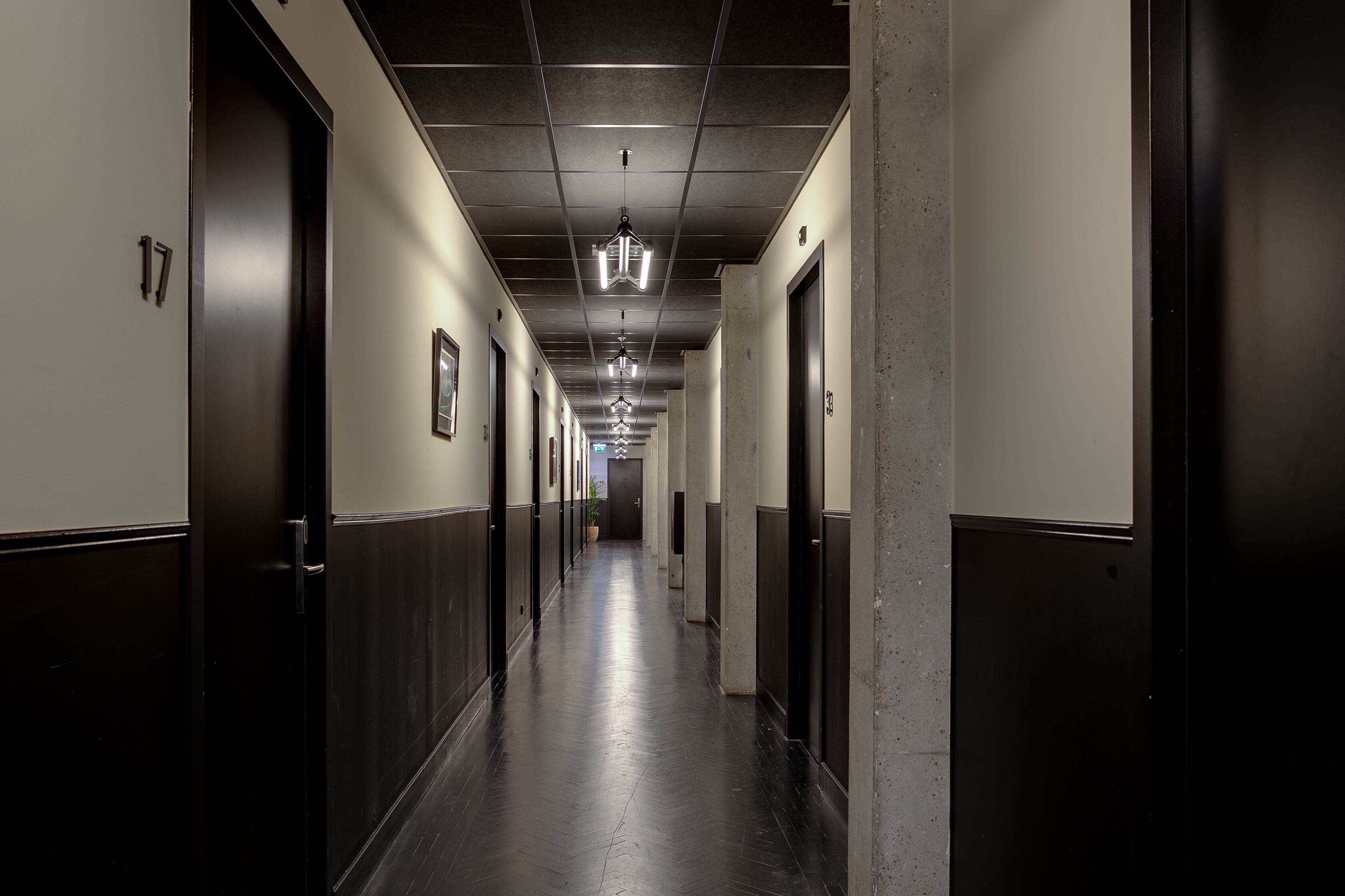 Een lange, smalle hotelgang met deuren aan beide kanten, genummerde bordjes en een centraal lichtarmatuur.