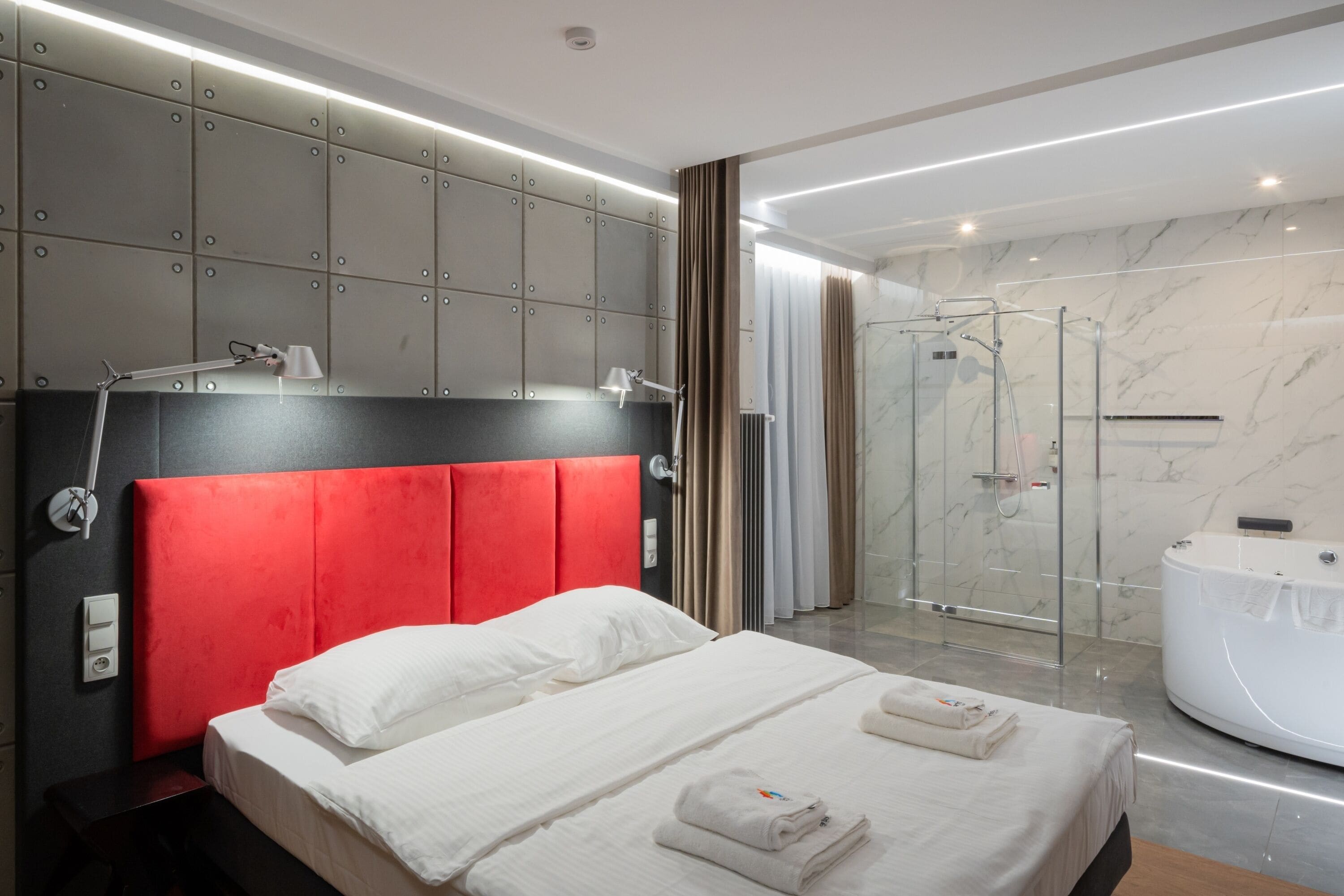 A modern hotel room with a red headboard, a large bed, a glass shower enclosure, and a white bathtub.