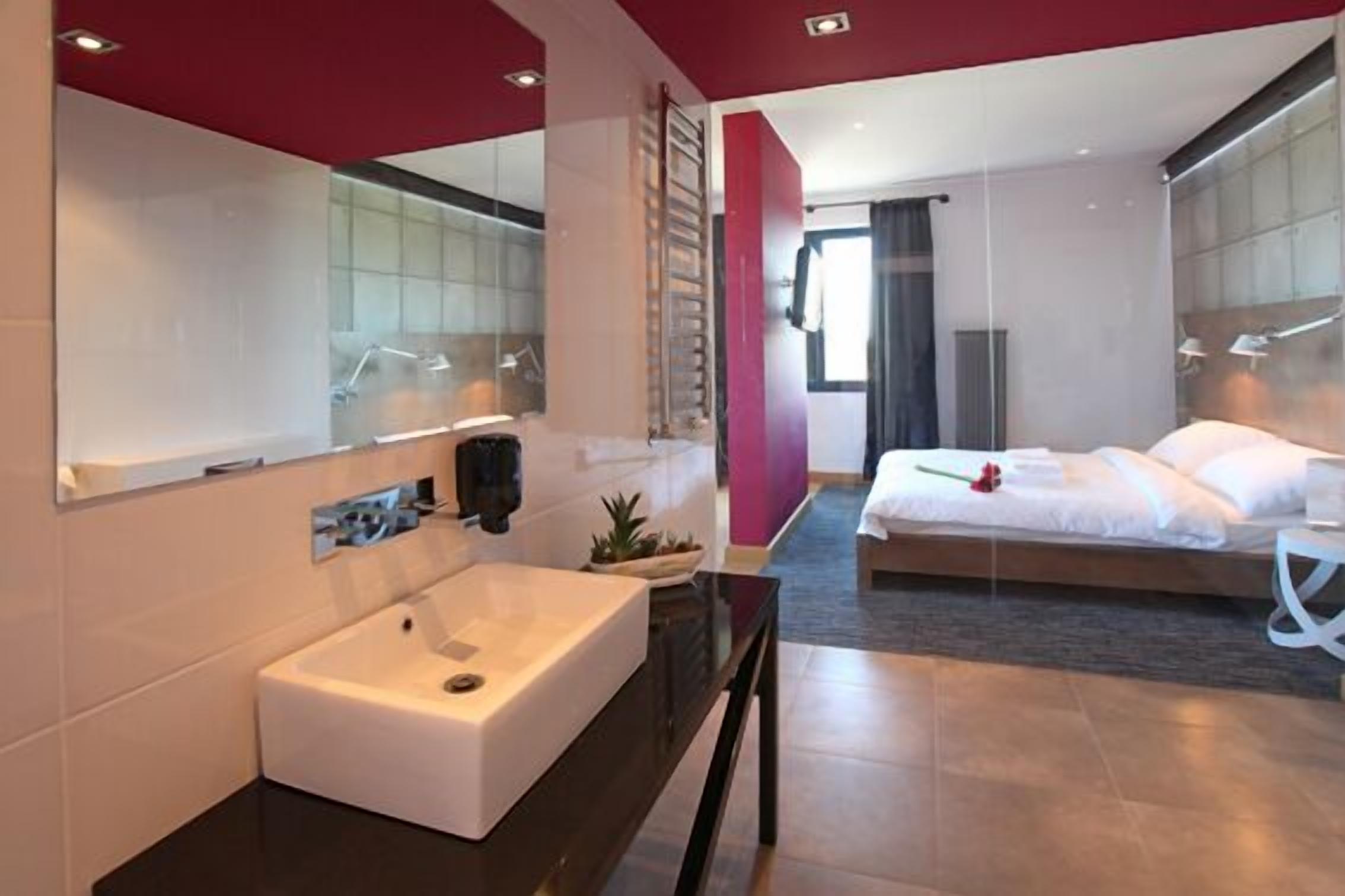 A modern hotel room with a large mirror, a white sink, a bed with white linens, and a small potted plant.