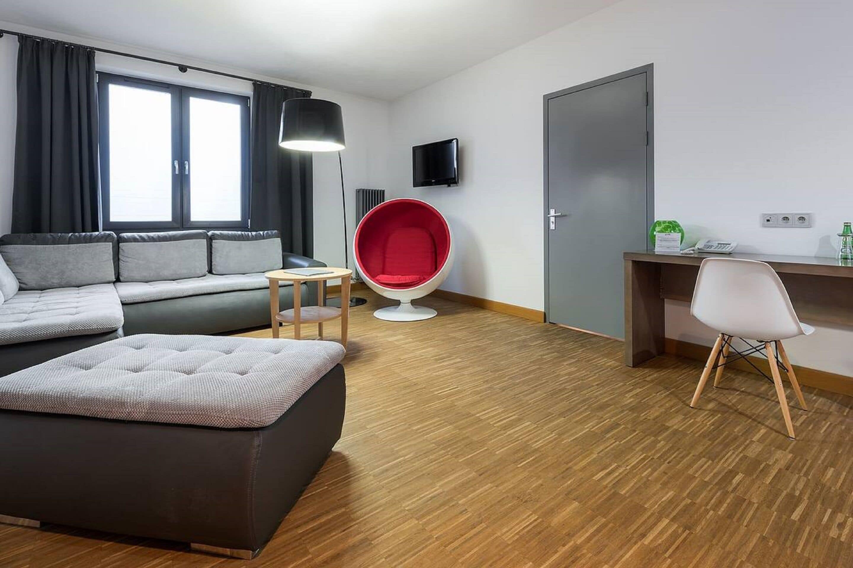 A modern room with a sofa, ottoman, red chair, desk, and a TV on the wall.