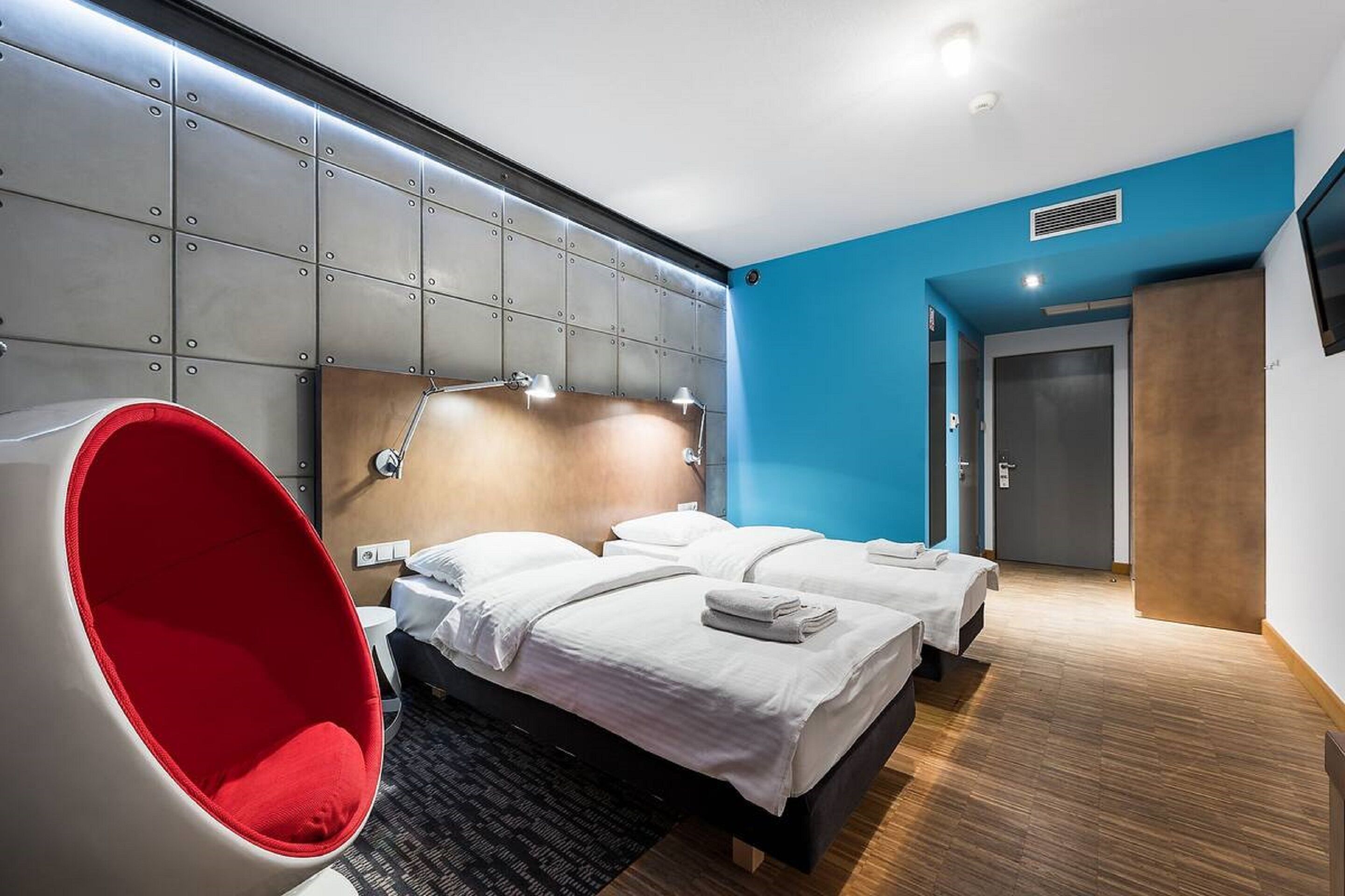 Una habitación de hotel moderna con una cama grande, una silla roja y una pared azul decorativa.