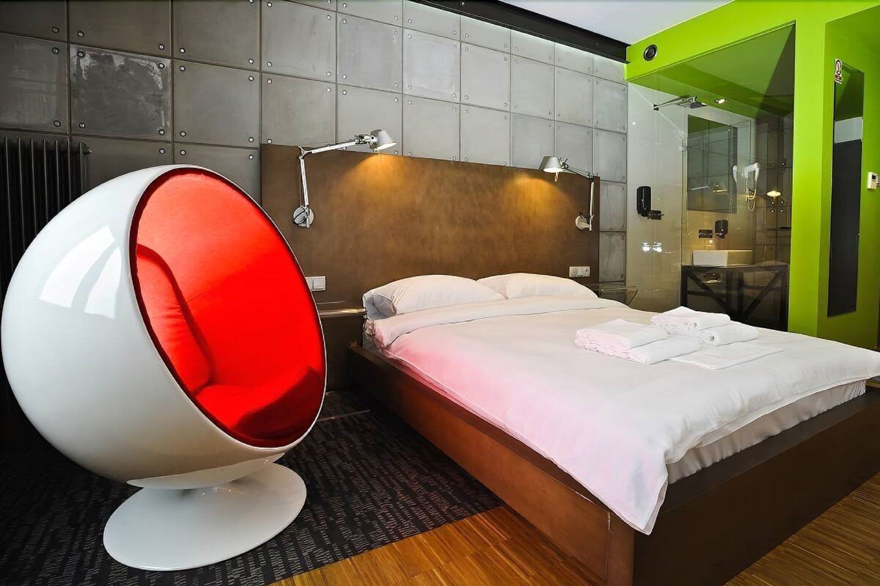 A modern hotel room with a large bed, a unique red egg chair, a wooden headboard, and a green accent wall.