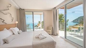 A modern hotel room with a large bed, a balcony with a table and chairs, and a scenic view of the sea.