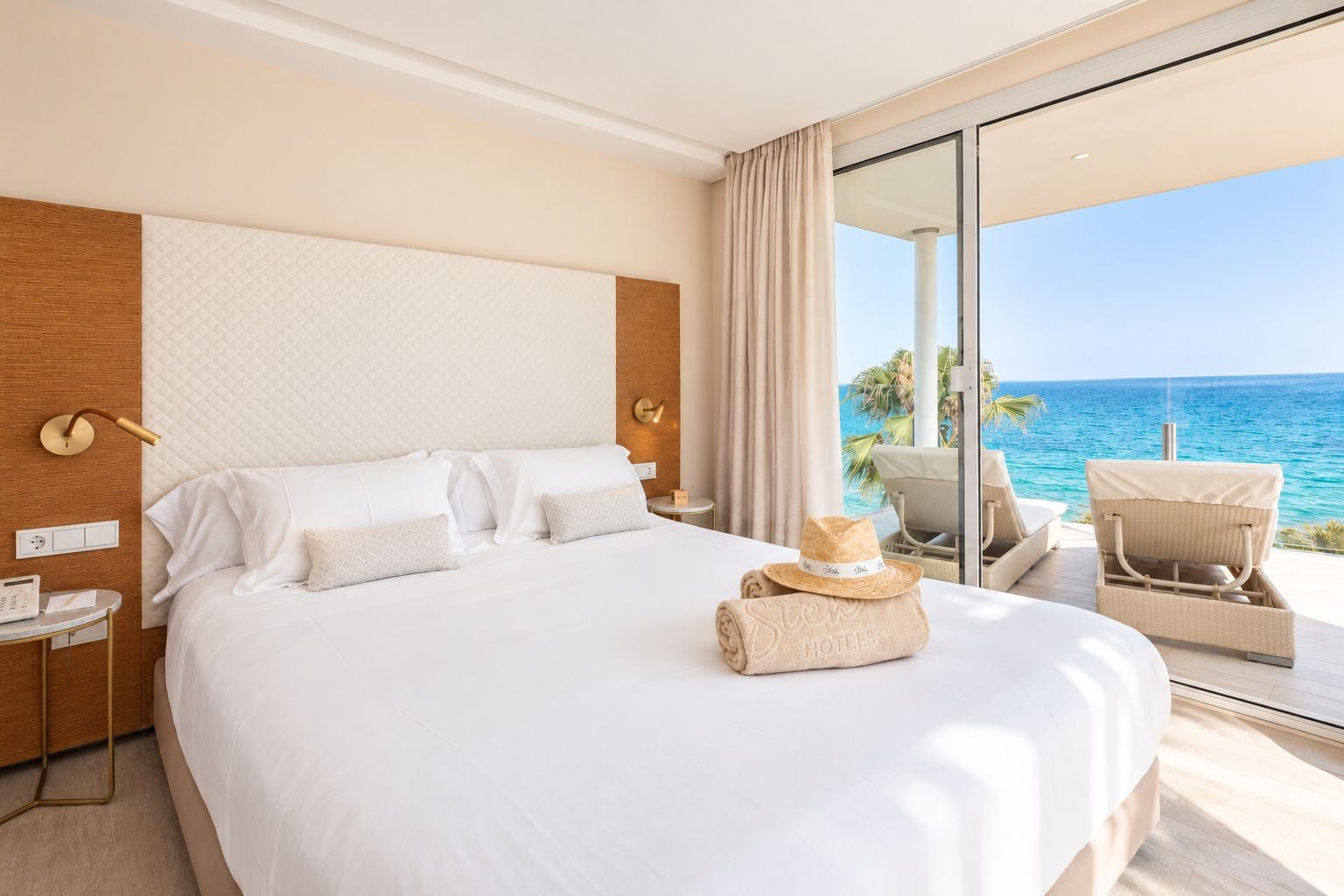 Une chambre d’hôtel avec un grand lit, une table de chevet avec une radio, une chaise près de la fenêtre et une vue sur l’océan.