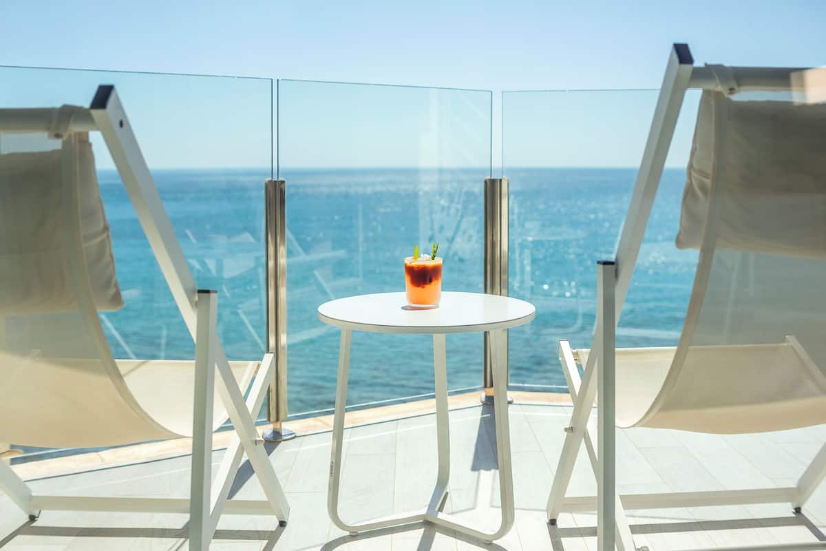 A balcony with two white lounge chairs, a small round table, and a glass of drink. The view includes a body of water and a distant cityscape.