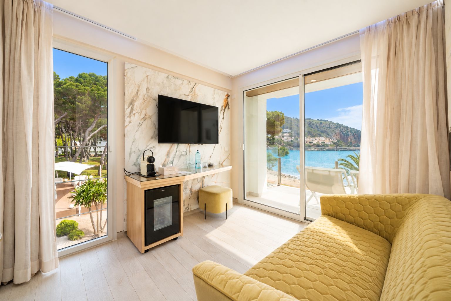 A hotel room with a large window, a flat-screen TV, a minibar, a yellow sofa, and a view of the sea.