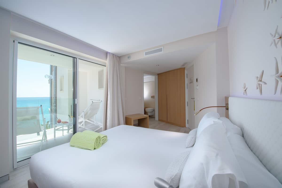 A hotel room with a large bed, a balcony with a view of the sea, a small bathroom, and a wooden wardrobe.