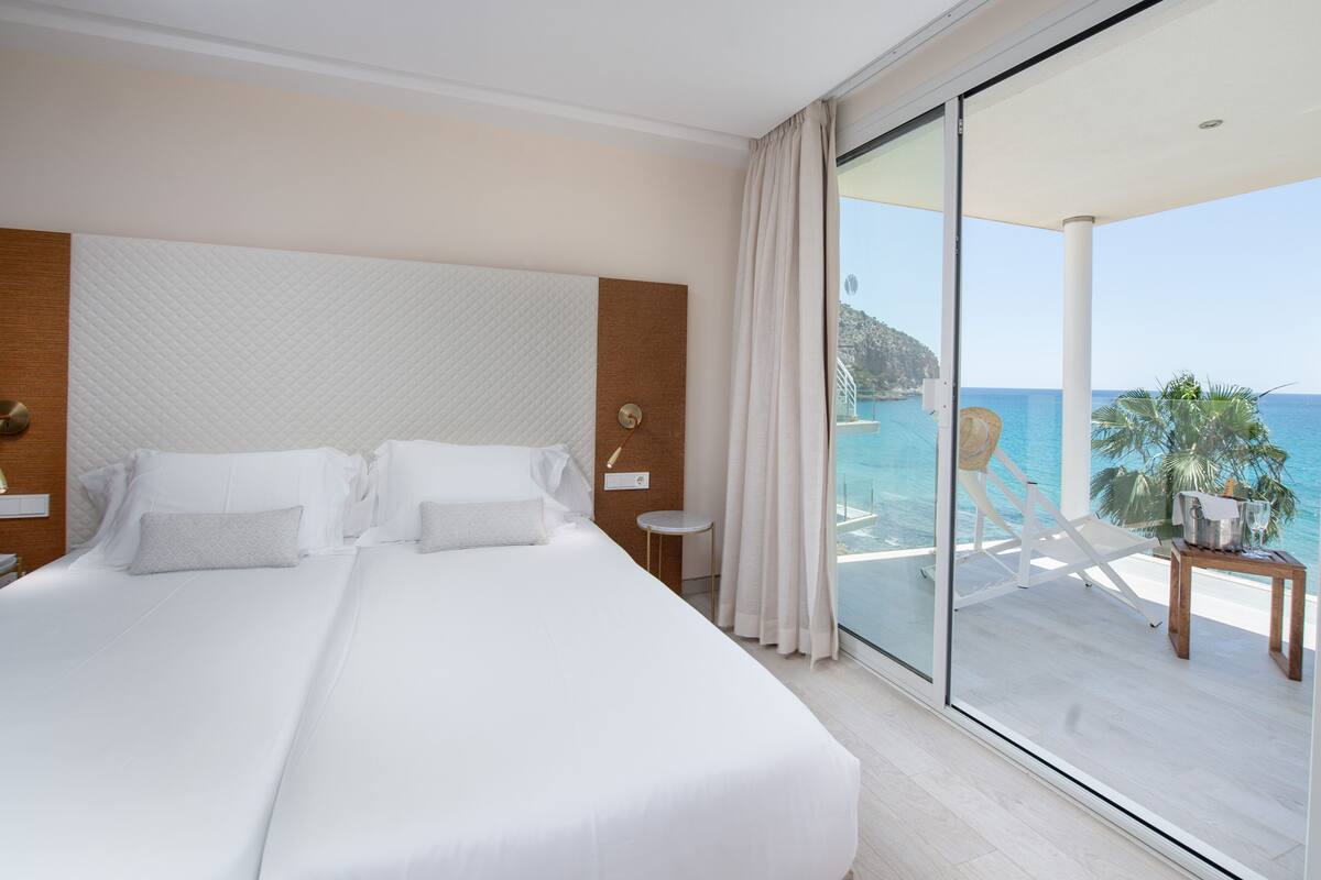 A hotel room with a large bed, a view of the sea, a balcony with a table and chairs, and a wooden headboard.
