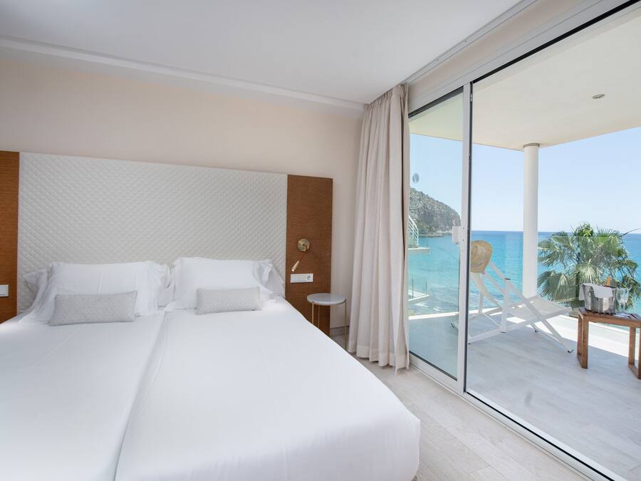 A hotel room with a large bed, a view of the sea, a balcony with a table and chairs, and a wooden headboard.