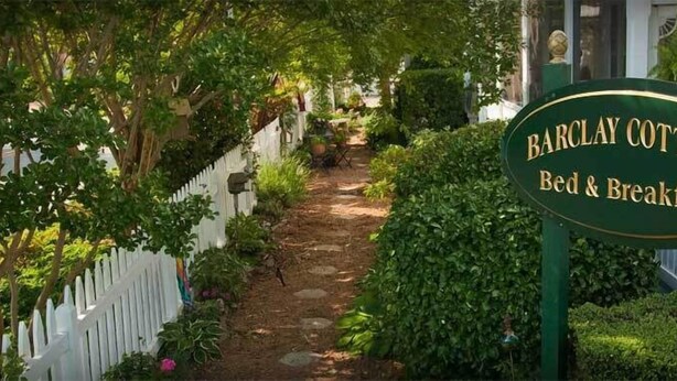 A sign for Barclay Cottage Bed & Breakfast, surrounded by lush greenery and a white picket fence. - Barclay Cottage (Virginia Beach)
