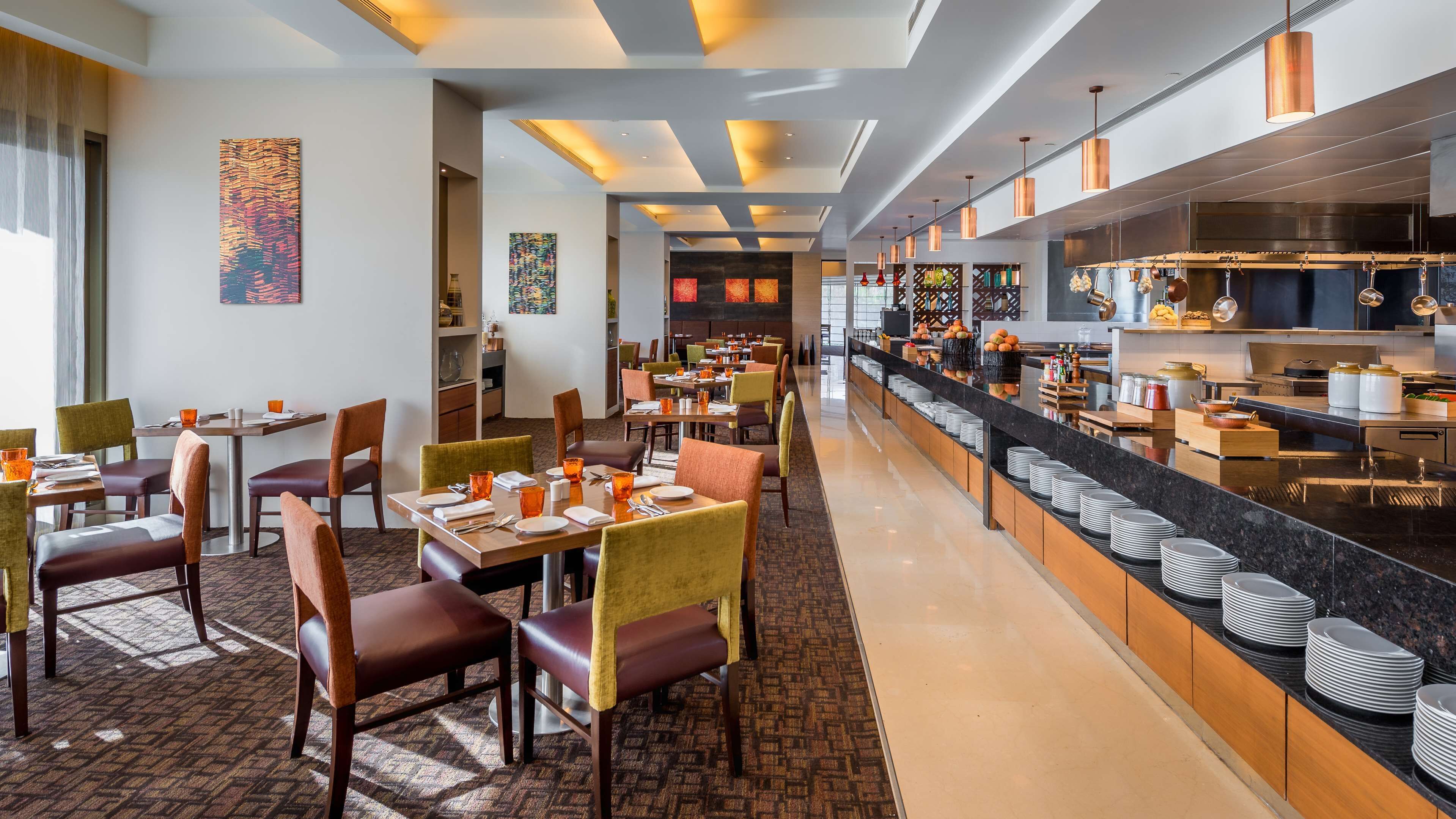 Un restaurant moderne doté de nombreuses tables et chaises, d’un long comptoir avec un buffet, et d’une cuisine visible au fond de la salle.