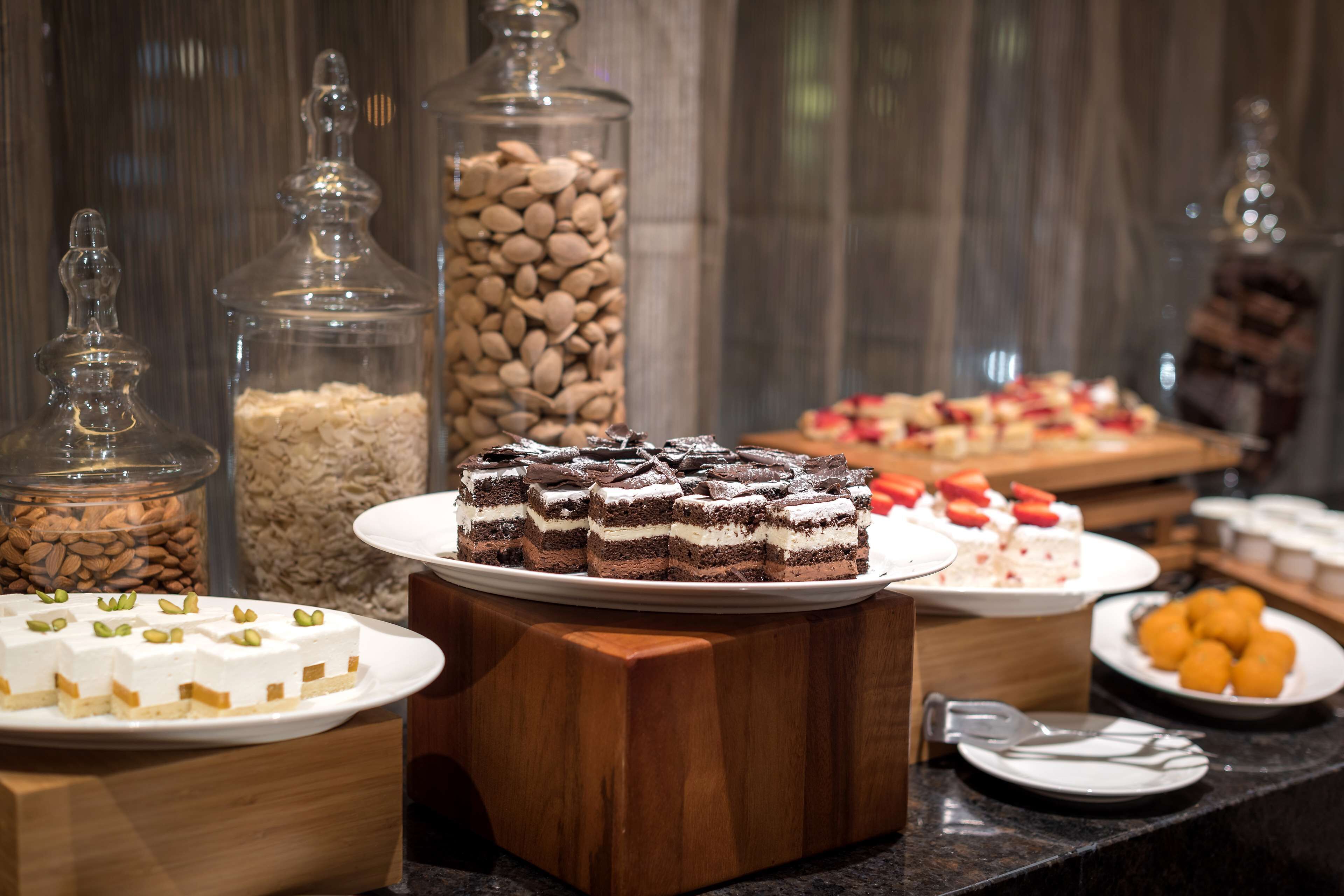 Ein Buffet mit verschiedenen Desserts, darunter Kuchen und Gebäck, präsentiert auf einem Holztisch. Im Hintergrund stehen durchsichtige Glasbehälter mit Nüssen und weiteren Speisen.