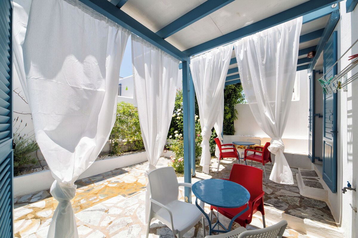 Un espace extérieur abrité avec des chaises blanches et rouges, une table bleue et des rideaux blancs.