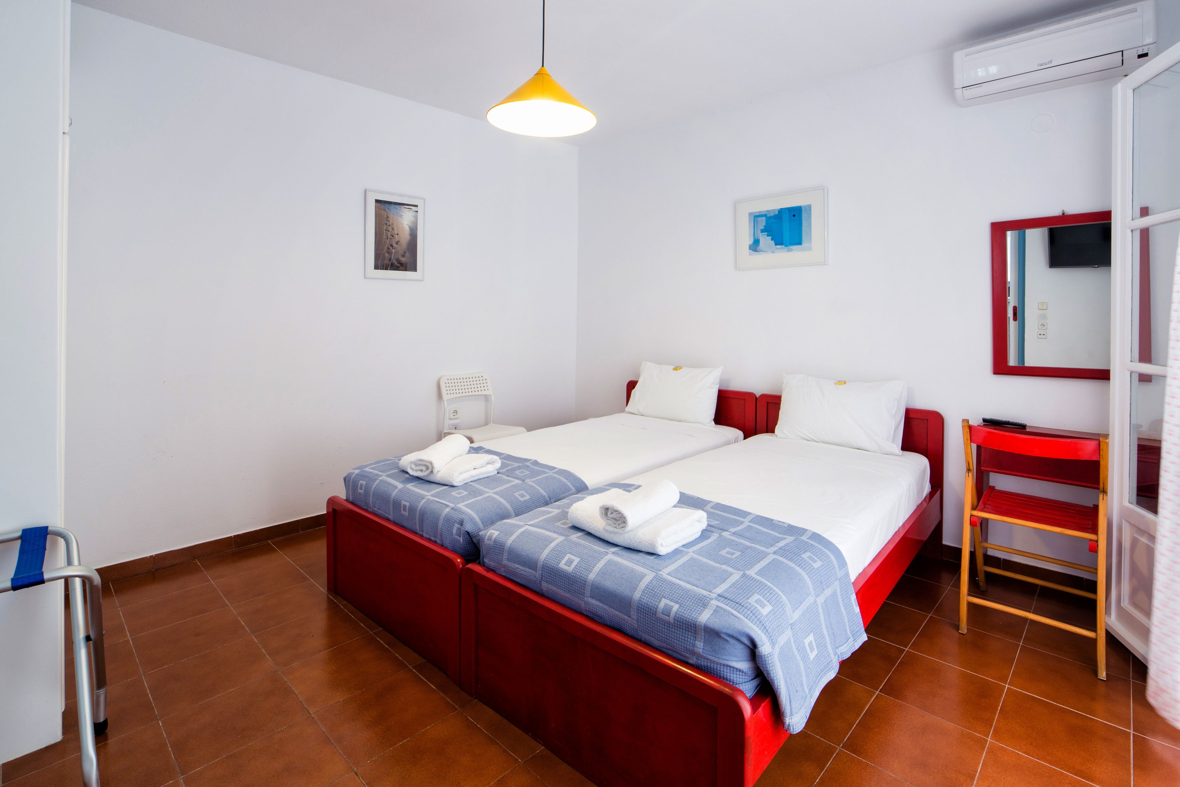 A hotel room with two beds, a red bench, a small table, and a wall-mounted air conditioner.