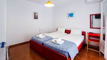 A hotel room with two beds, a red bench, a small table, and a wall-mounted air conditioner.