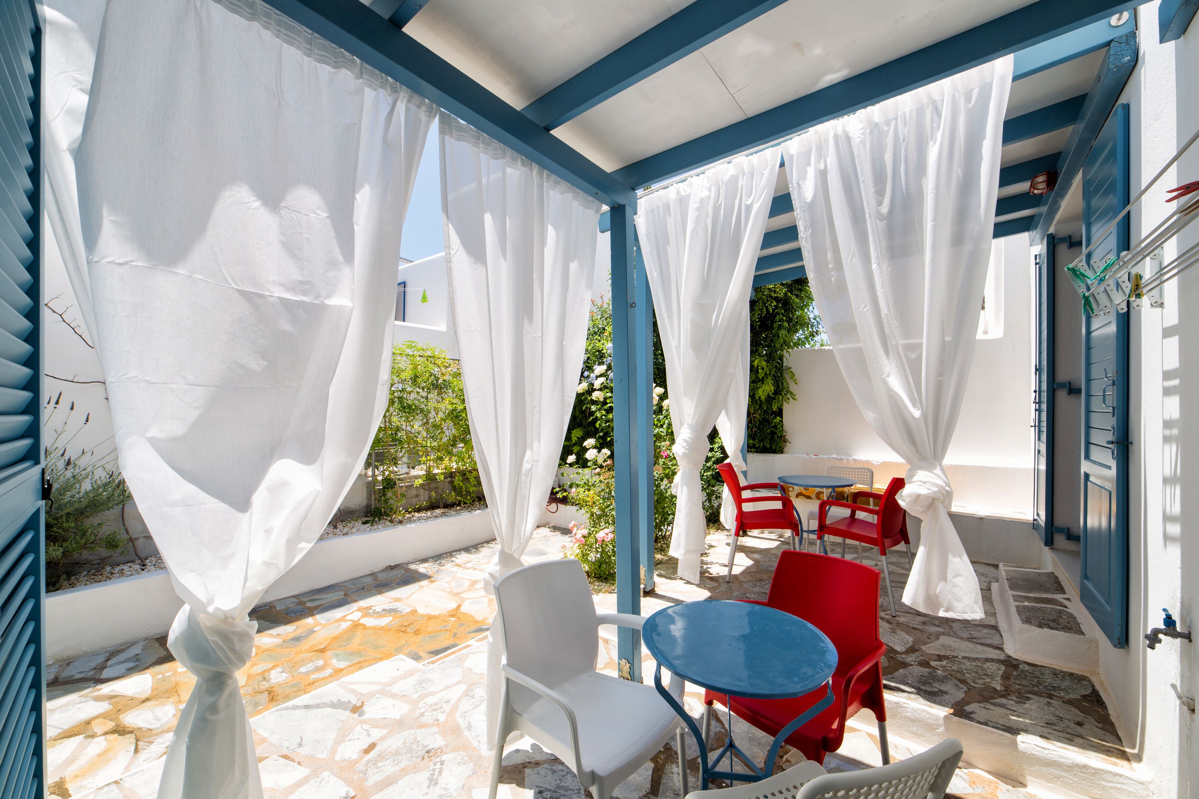 A covered outdoor seating area with white and red chairs, a blue table, and white curtains.