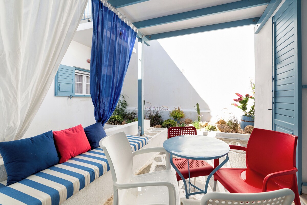 A covered outdoor seating area with a white sofa, red and blue cushions, a small round table, and a red chair.