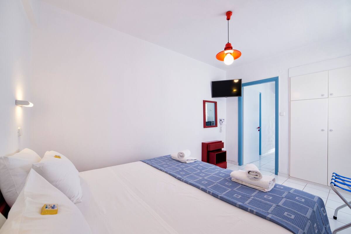 Una habitación de hotel con una cama, una lámpara colgante roja, un televisor, una tocador rojo, una puerta azul y un armario blanco.