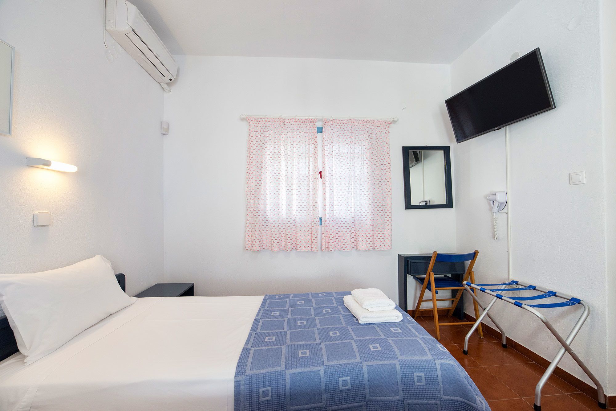 Ein Hotelzimmer mit Bett, Wandfernseher, Fenster mit Vorhang, Stuhl und kleinem Tisch.