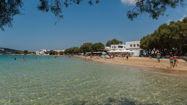 A beach with clear water, people swimming, and buildings in the background. - Fivos Apartments (Paros)